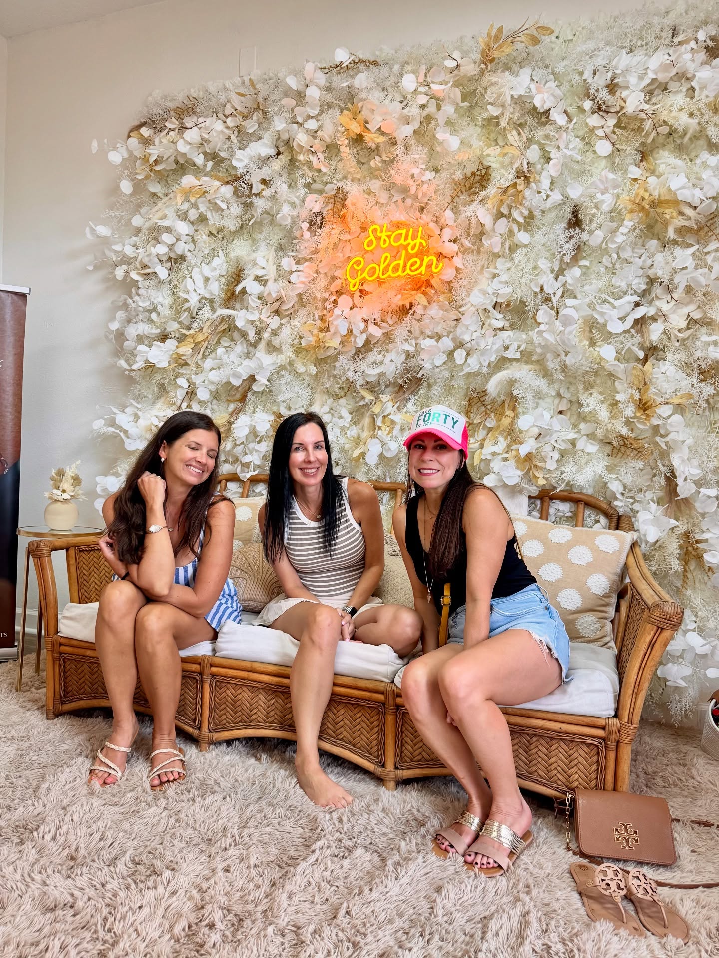 Three women sitting on a wicker bench in front of a decorative wall with 'Stay Golden' sign.
